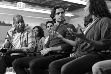 A group of adults are shown seated, having a discussion in a town hall setting.