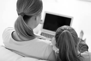 A mom and daughter are seated next to each other on a couch, looking at a laptop screen.