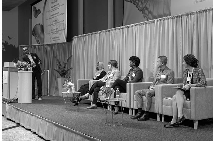 Dr. Bignall is standing at a podium, facing the five panelists who are seated on the stage.
