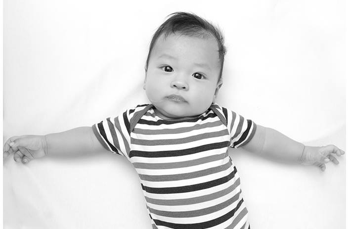 A young infant is laying down with arms sprawled out. The infant is wearing a striped onesie.