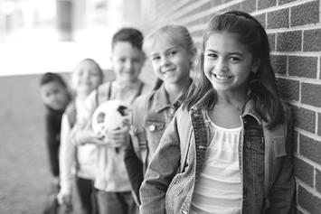 A group of five elementary aged kids are standing against a brick wall. They are outdoors. One is holding a soccer ball.