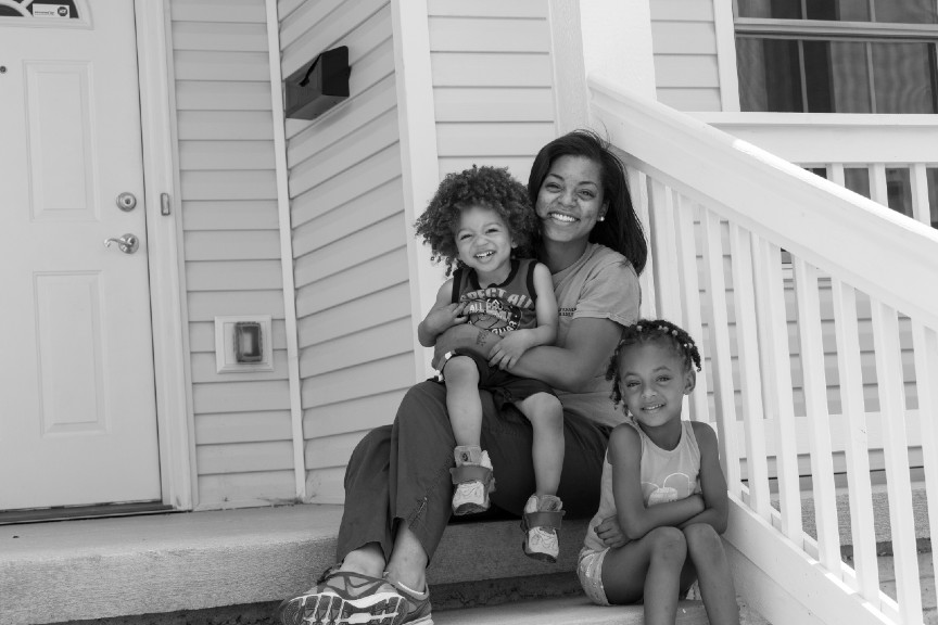 A mom is holding one child while sitting on her front porch, and her other child is sitting next to her. 