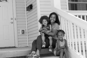 A mom is holding one child while sitting on her front porch, and her other child is sitting next to her.