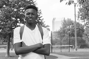 A smiling teenage boy is wearing a backpack and standing in a park. His arms are crossed.