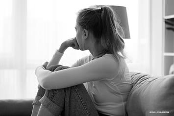 A concerned teen girl sits on the couch, solemnly looking toward the window