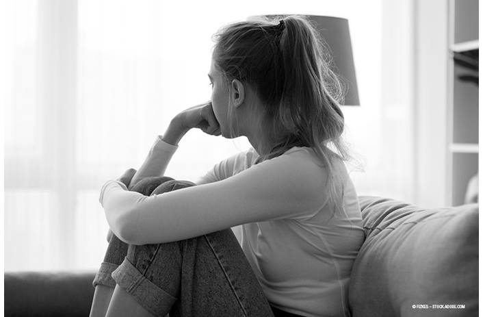 A concerned teen girl sits on the couch, solemnly looking toward the window