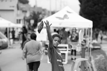 A boy is playing corn hole during a PlayStreets event. People behind him are gathering and playing in the street.