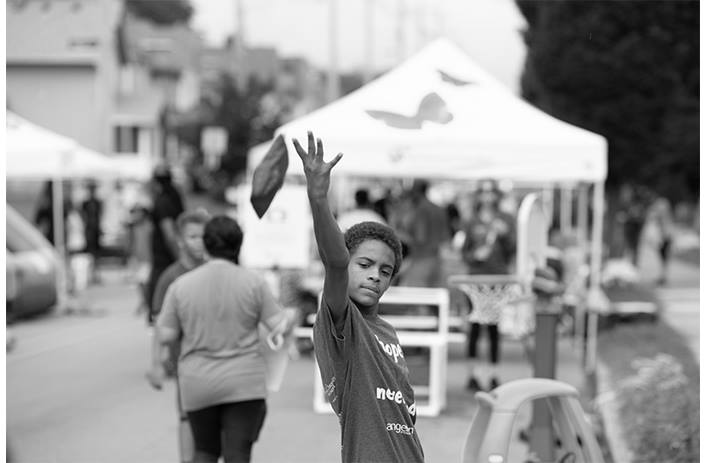 A boy is playing corn hole during a PlayStreets event. People behind him are gathering and playing in the street.
