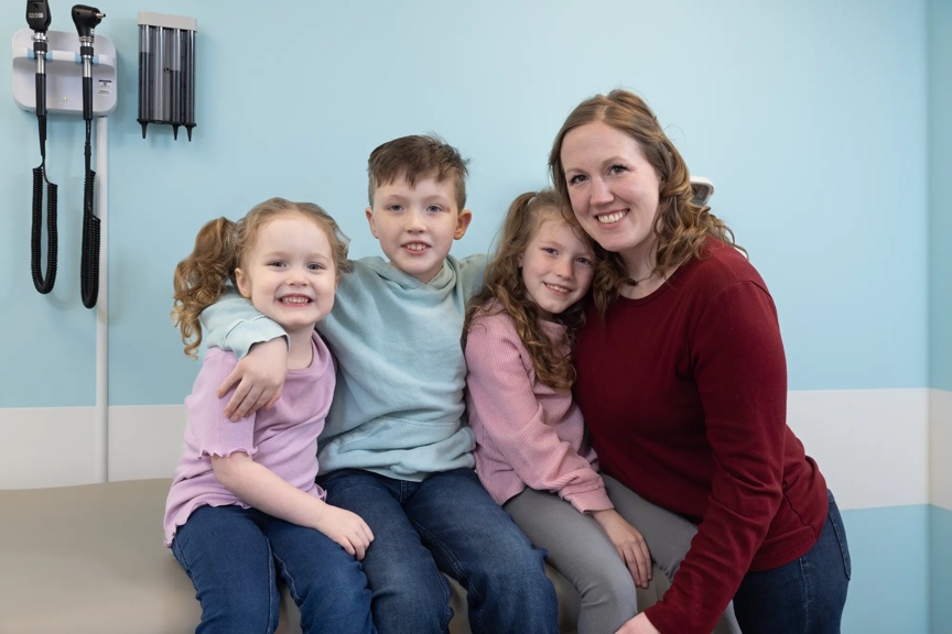 A smiling mom stands next to her three children, seated in a bright blue-walled clinic room.