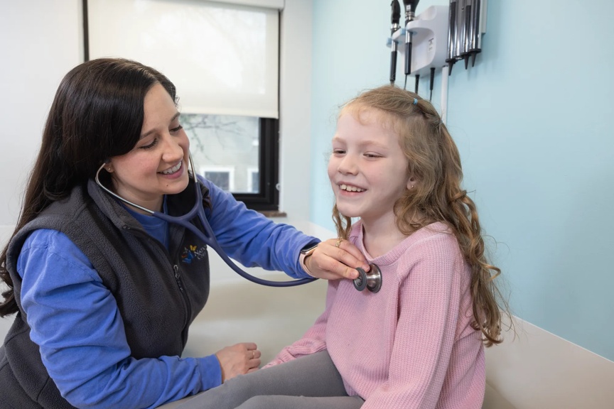 A clinician smiles while placing a stethoscope on a smiling patient in a clinical setting.