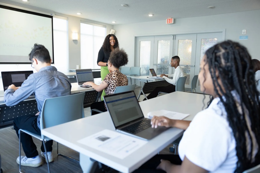 Several Boost program participants are seated, with computers, in a bright classroom-like setting.