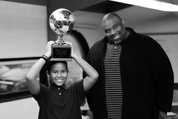 Tekle is smiling, holding the soccer championship trophy over his head. Wes is standing next to him, smiling.