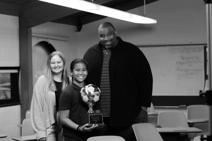 Taylor, Tekle and Wes are standing in a classroom. All are smiling. Tekle is holding a trophy with a large soccer ball on it.