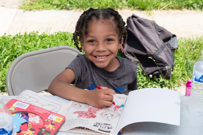 A smiling preschool aged child is seated at a table, coloring. A sidewalk and grass are in the background.