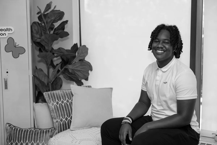 Ma'Kye is seated on a couch, smiling, wearing a white polo.