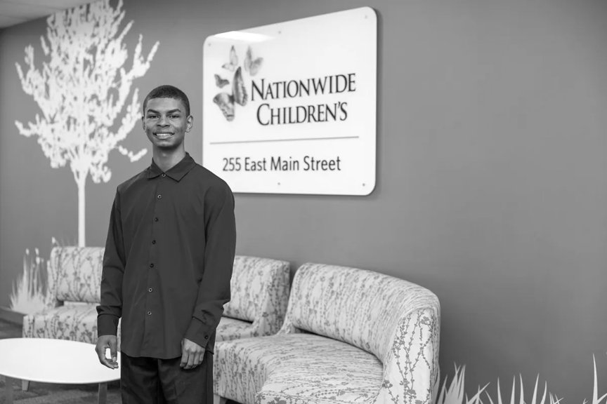 Ja’Marre is wearing a long-sleeved button down shirt, smiling, standing in front of a Nationwide Children's sign in an office entryway.