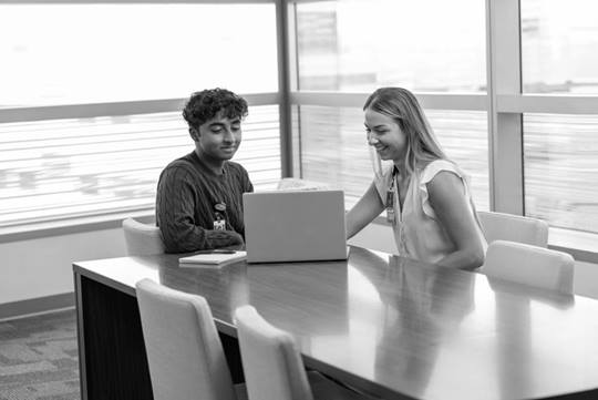 A mentor sits with her mentee at a table. They are looking at her laptop, smiling. A bright wall of windows is behind them. 