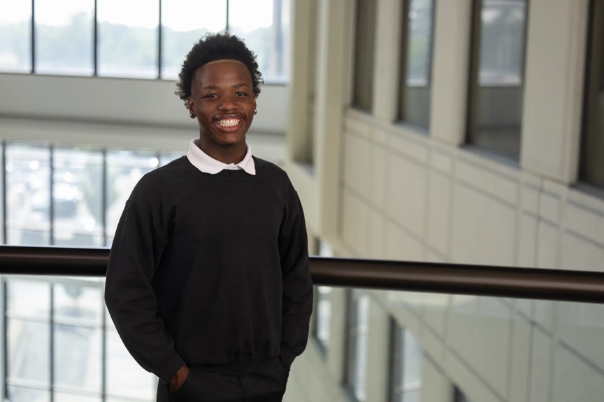Micheal smiles, wearing all black while standing in an office building with a wall of windows behind him.