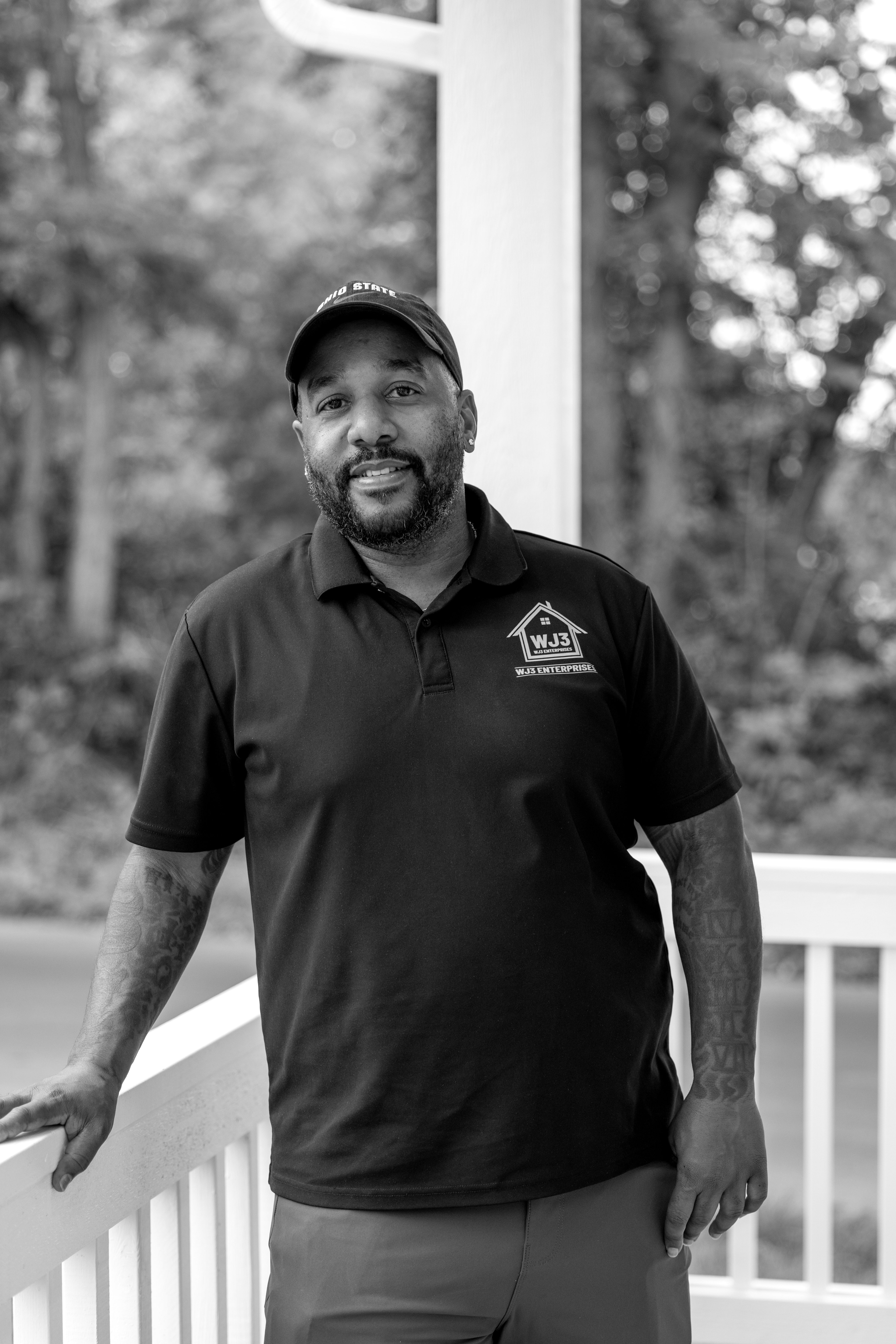 Wade wears a dark short sleeve polo with his business logo on it. He's smiling, resting his arm on a front porch railing.