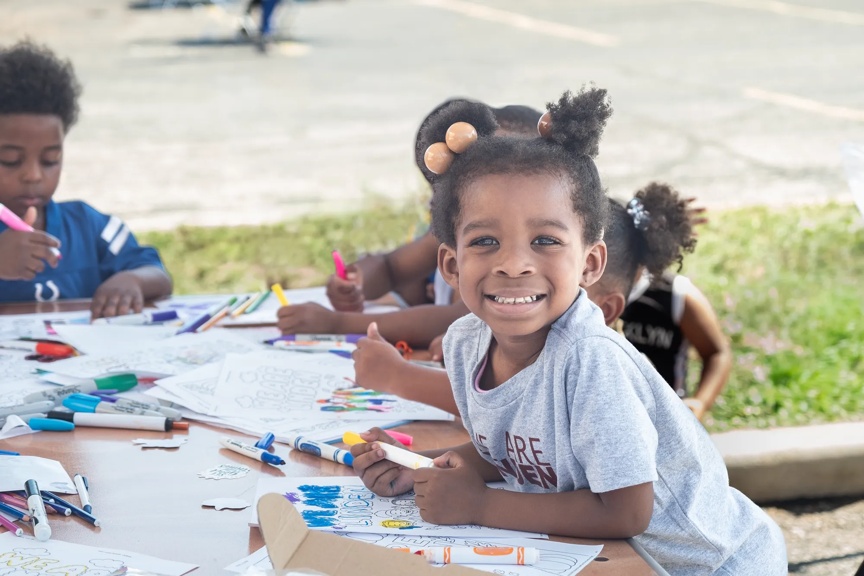 A smiling young kid is coloring at an outdoor table with other kids in the background.