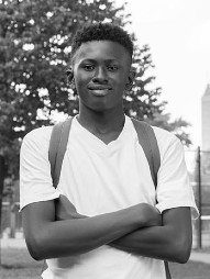 A high school boy stands outside with arms crossed, grinning. He's wearing a back pack.