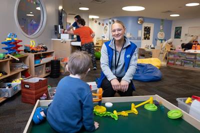 A hospital volunteer sitting and engaging with a young child playing at a table in a colorful playroom, with toys and other children in the background.