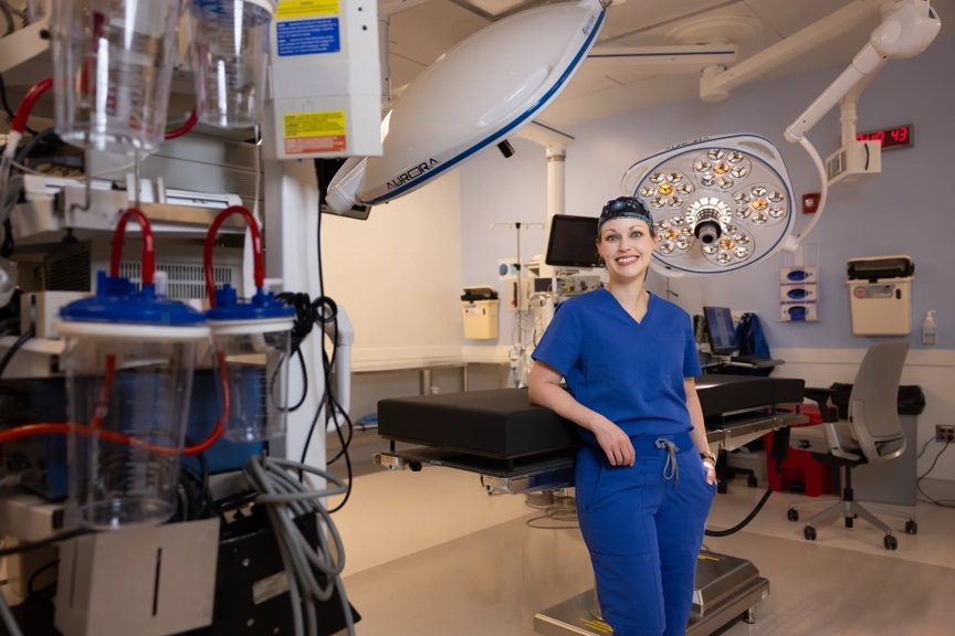 Dr. Stephanie Russo is a hand and upper extremity surgeon featured in this photo. She is in her blue scrubs with one hand in her pocket and the other at her side while standing in the operating room.