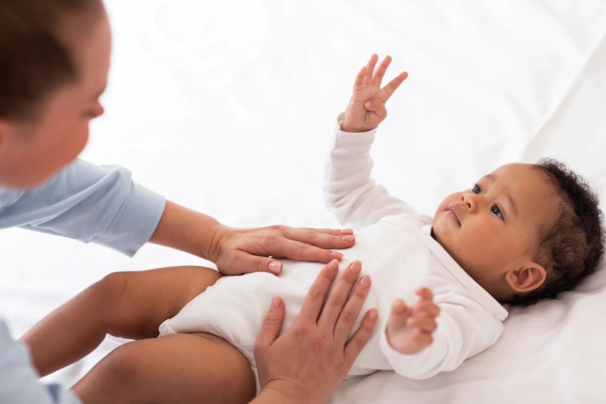 Infant in white onesie lays on back, with a caregiver gently touching the baby's stomach.