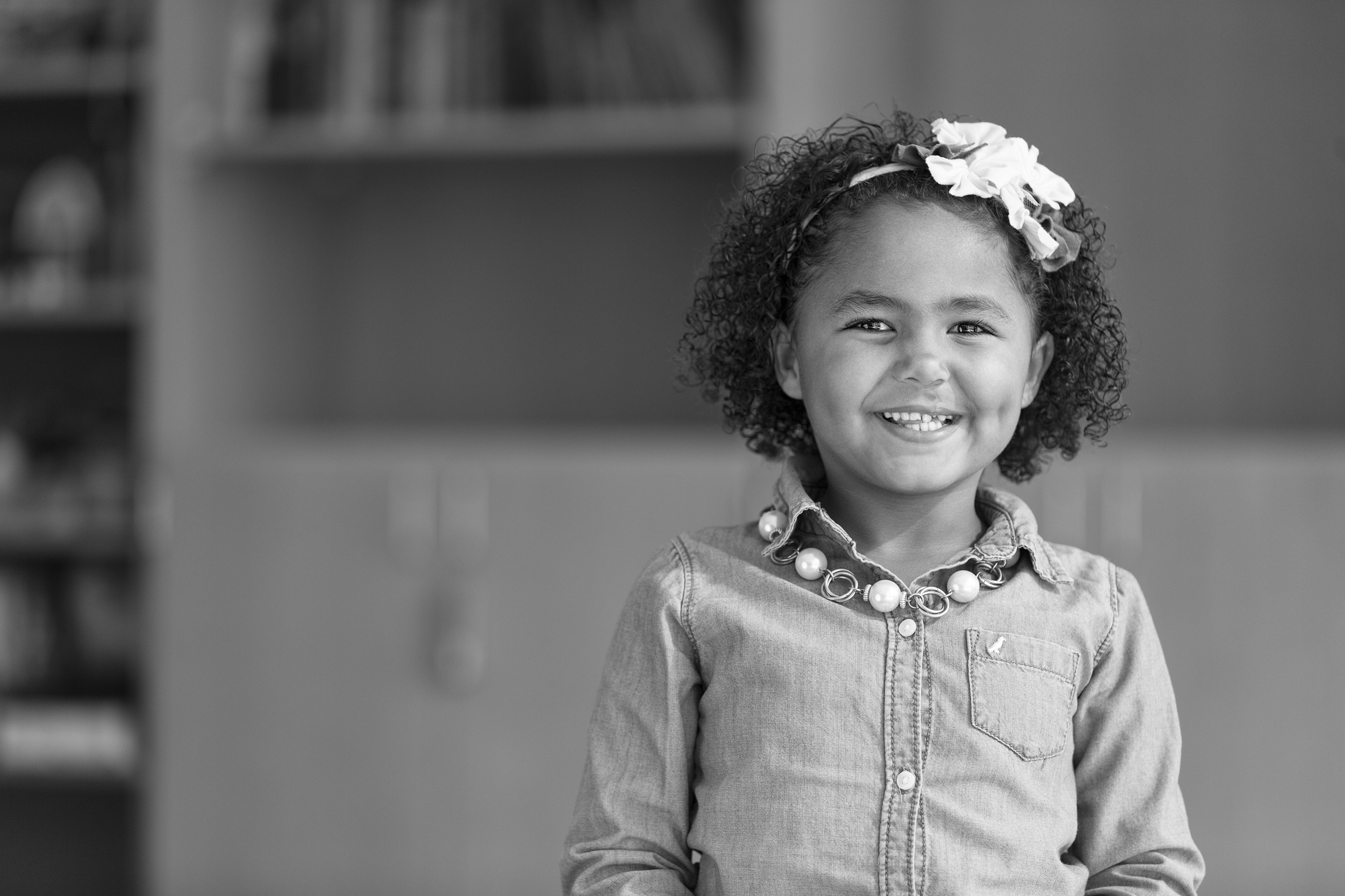 A young child is standing indoors wearing a long-sleeve button-up shirt with a front pocket and a necklace made of large beads. The child has curly hair adorned with a large flower headband. In the background, there are shelves with books and cabinets, suggesting a classroom or learning environment.