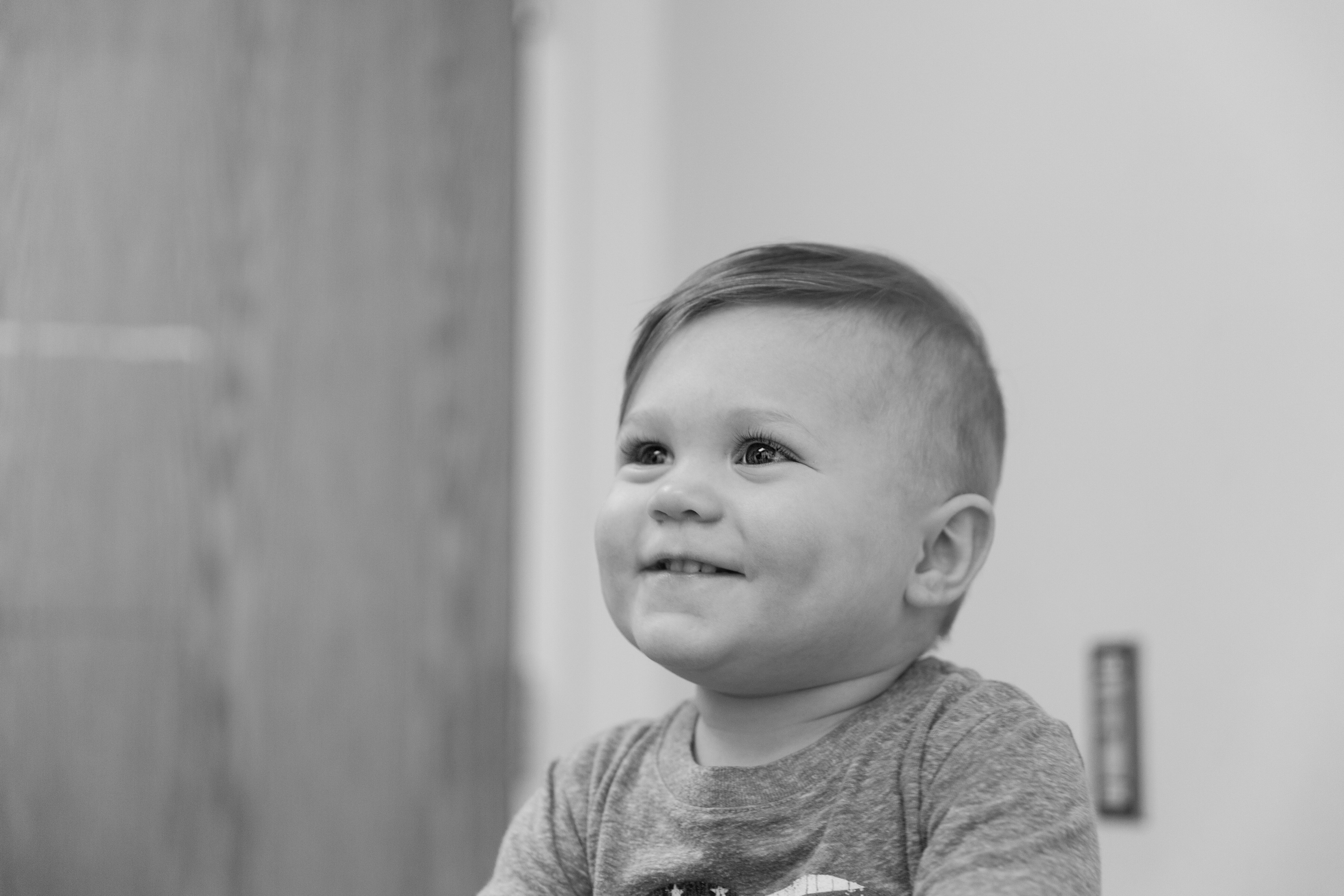 Black-and-white photo of a young child wearing a short-sleeved shirt with a graphic design, sitting indoors. A wooden door is visible in the background on the left, and a light-colored wall with a rectangular object, possibly a switch plate, is on the right. The child’s hair is neatly combed to the side.
