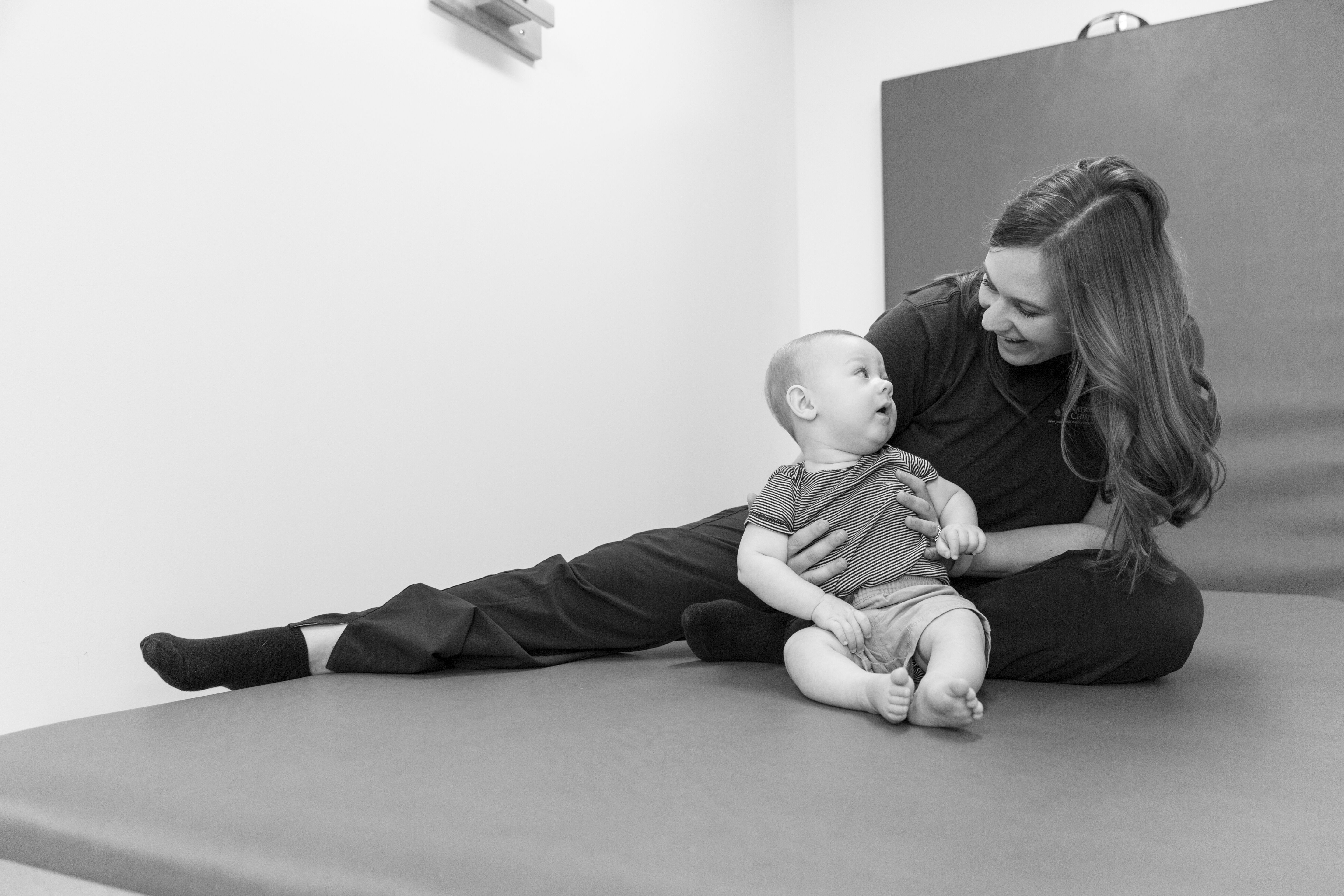 An adult is sitting on a padded mat in a therapy or playroom, with legs extended to the side, supporting a baby who is sitting upright. The baby is wearing a striped shirt and shorts, and the adult is dressed in dark clothing. The background includes a plain wall and a large cushioned panel.