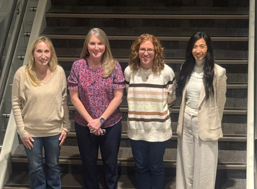 Four individuals standing side by side at the bottom of a wide staircase with metal railings on both sides. The group is positioned indoors on a carpeted floor. Each person is dressed in casual or business-casual attire: the first person from the left wears a beige sweater with jeans and brown shoes; the second person wears a patterned short-sleeve top with dark pants and black shoes; the third person wears a white sweater with horizontal brown and gray stripes, dark pants, and white sneakers; the fourth person wears a light-colored suit with a white turtleneck and white shoes. The staircase behind them has dark steps and appears to lead to an upper level. Lighting is bright and even.