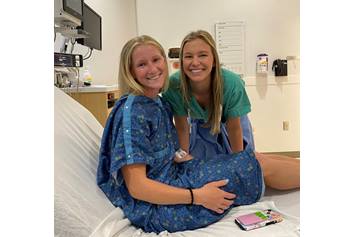 Bailey in a hospital gown sitting on a hospital bed smiling next to a nurse
