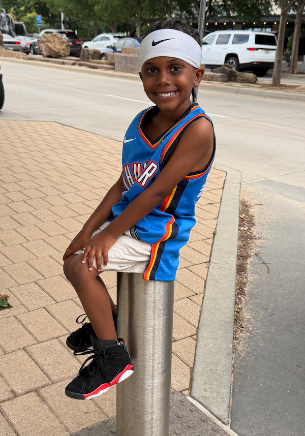 young boy sitting on sliver pole smiling wearing a basketball jersey