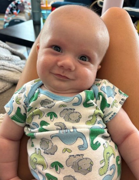 Baby Kace laying on a lap smiling wearing a dinosaur onesie