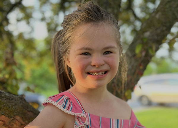 A smiling young girl stands outdoors beside a tree, wearing a pink striped dress with ruffled sleeves. She poses confidently with one hand on her hip in warm evening sunlight, with green grass and trees softly blurred in the background.