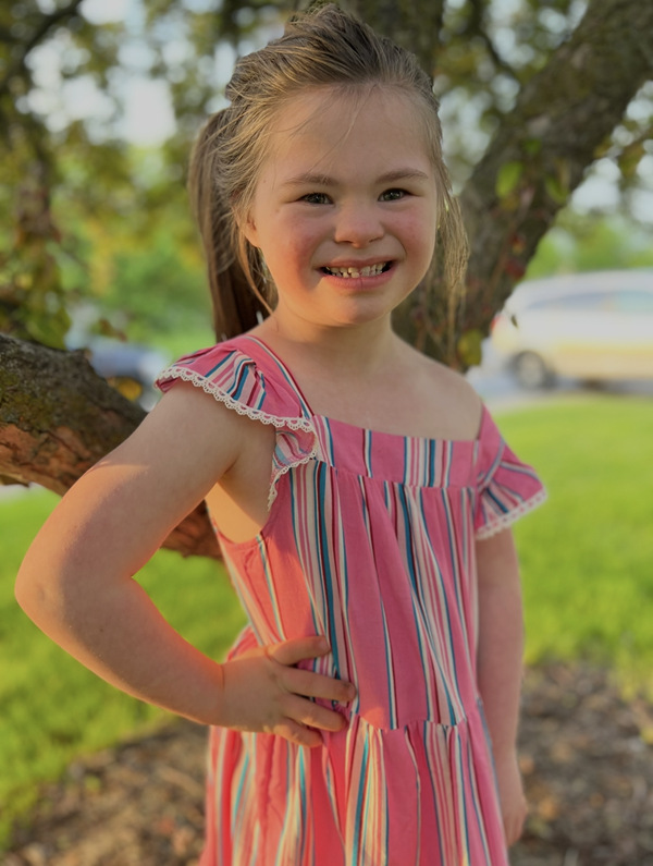 A smiling young girl stands outdoors beside a tree, wearing a pink striped dress with ruffled sleeves. She poses confidently with one hand on her hip in warm evening sunlight, with green grass and trees softly blurred in the background.