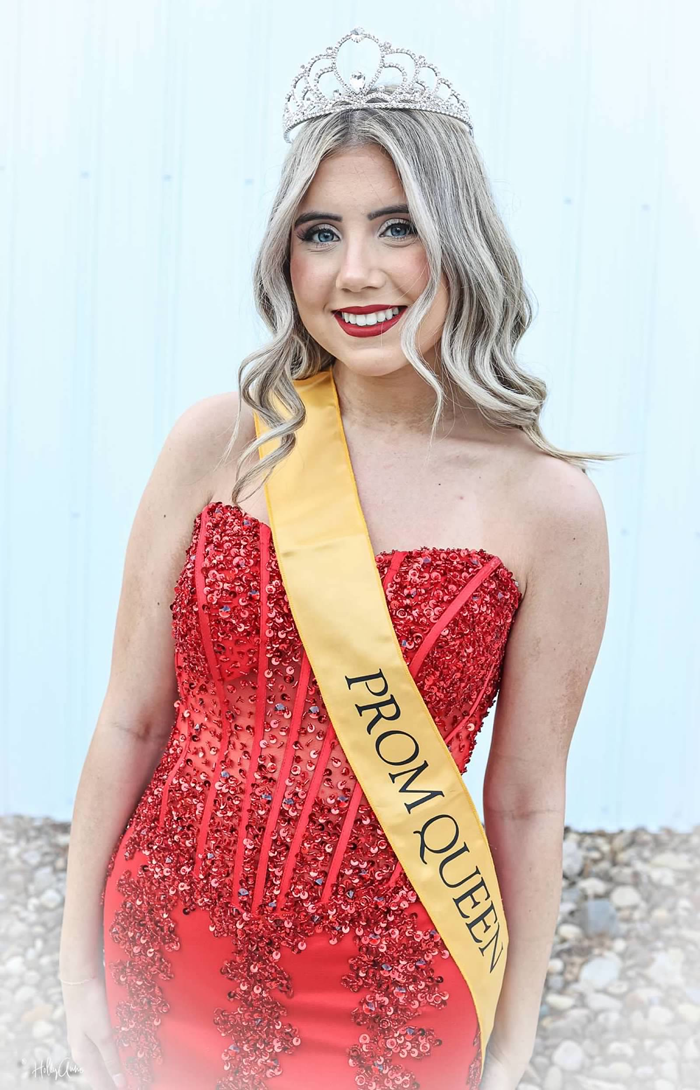 Macy stands smiling in a sparkly red strapless gown with a jeweled tiara on her head and a yellow sash that reads "Prom Queen." 