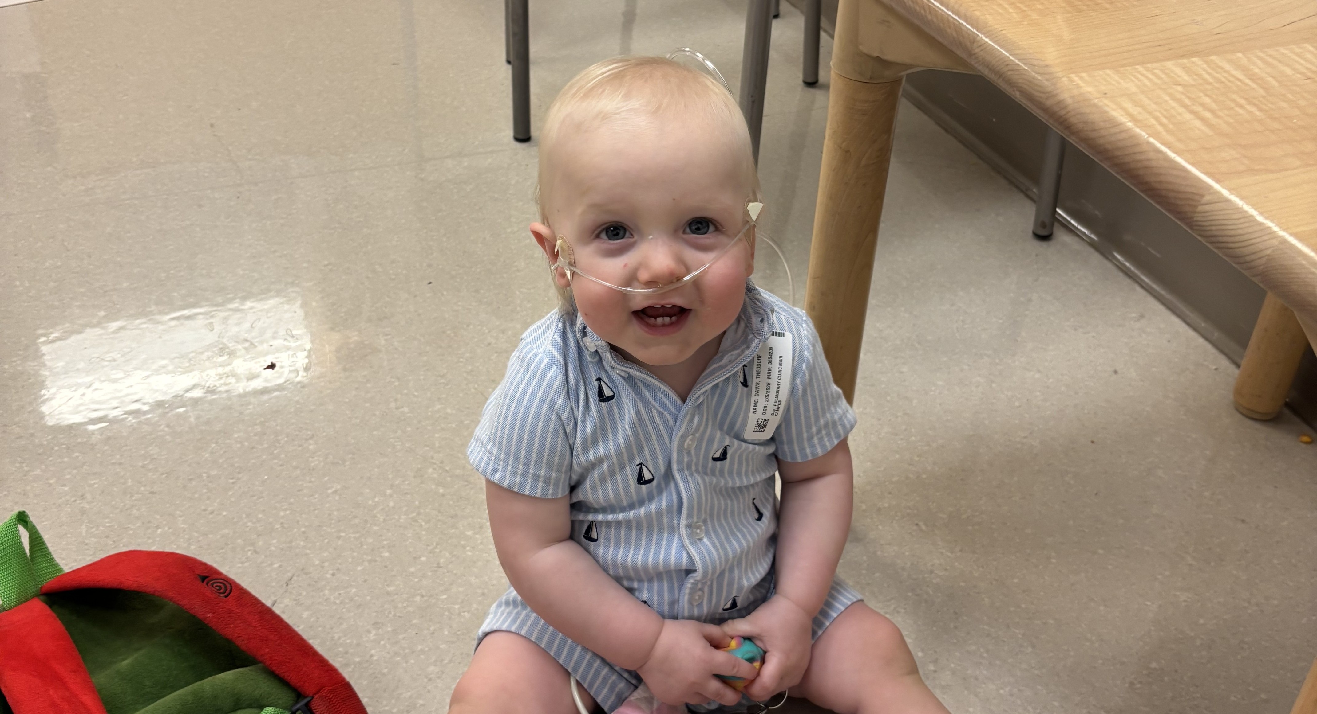 A smiling toddler sits on the floor of a clinic exam room holding a toy and a set of keys. The child wears a nasal cannula for oxygen support and is surrounded by a small backpack and medical tubing.