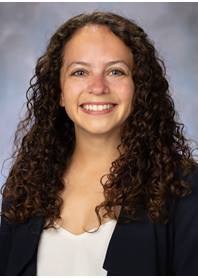 Portrait of Alex Winter. Alex has long, curly brown hair. She is wearing a black blazer and white top.