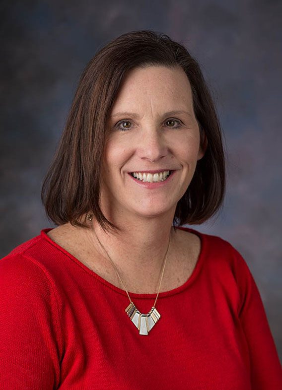 Portrait of Amy Donegan, PCPNP. Amy has short brown hair, a red shirt and silver necklace on. The background is a mix of dark and light grays. 