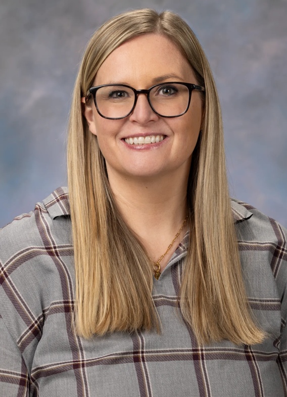 Portrait of Annie Gamble, woman with long blonde hair, glasses, and a patterned grey shirt.