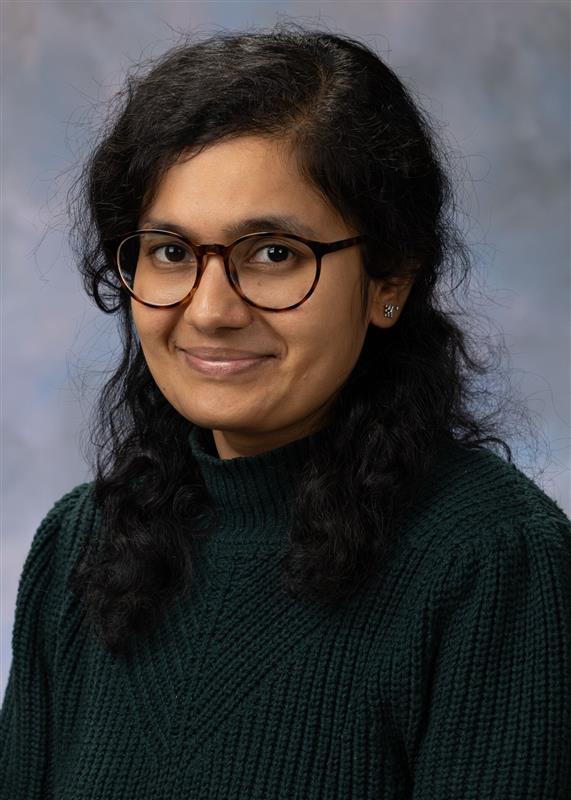 Portrait of Archana Mahadevan Iyer. Archana has long black hair and is wearing glasses and a black sweater. 