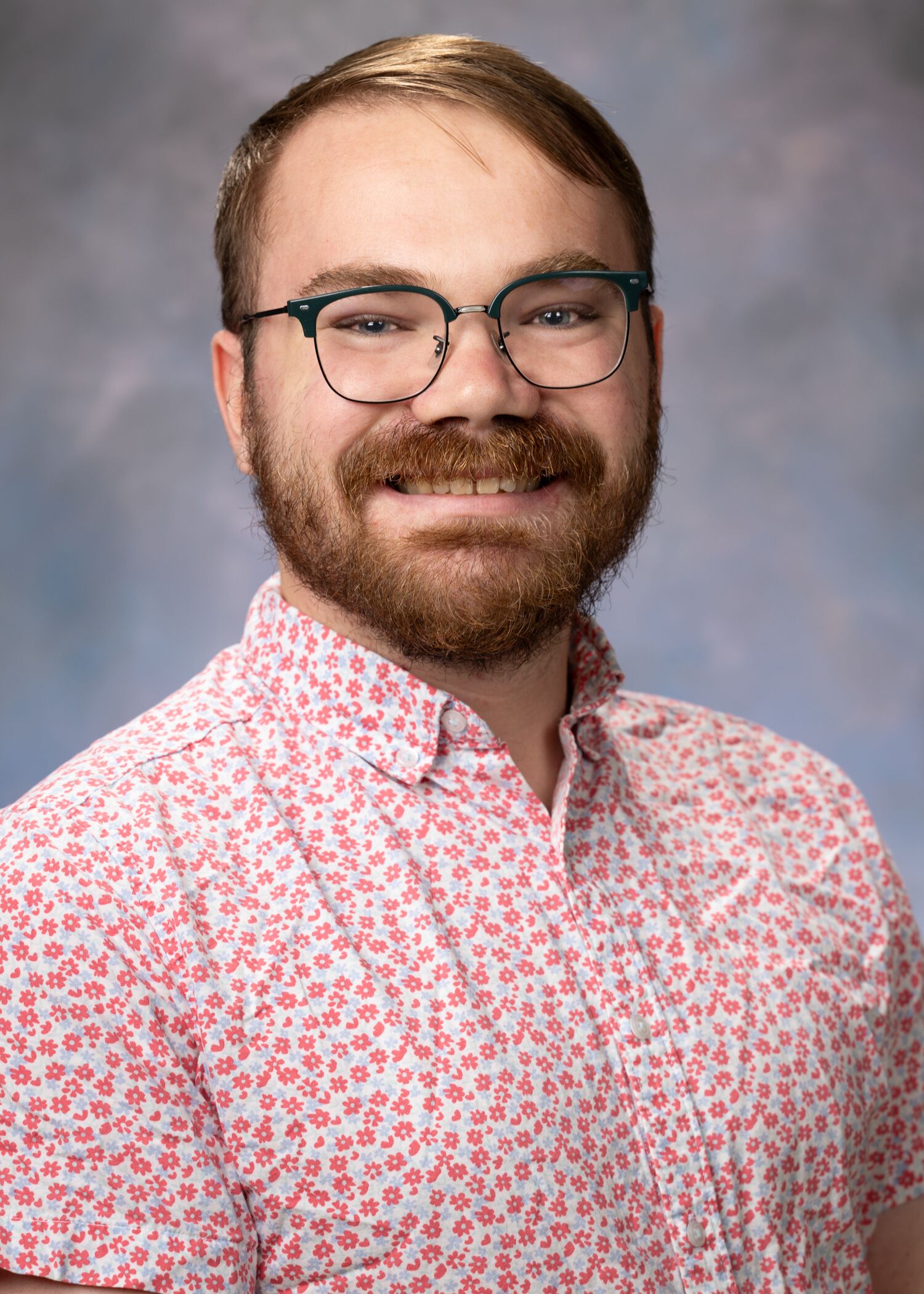 Portrait of Colin Maguire. Colin has short light brown hair with a beard. Colin is wearing glasses and a red and white spotted button-up shirt.