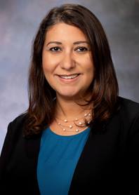 Portrait of Diana Bharucha-Goebel. She has shoulder length brown hair, a blue shirt and a black jacket on.