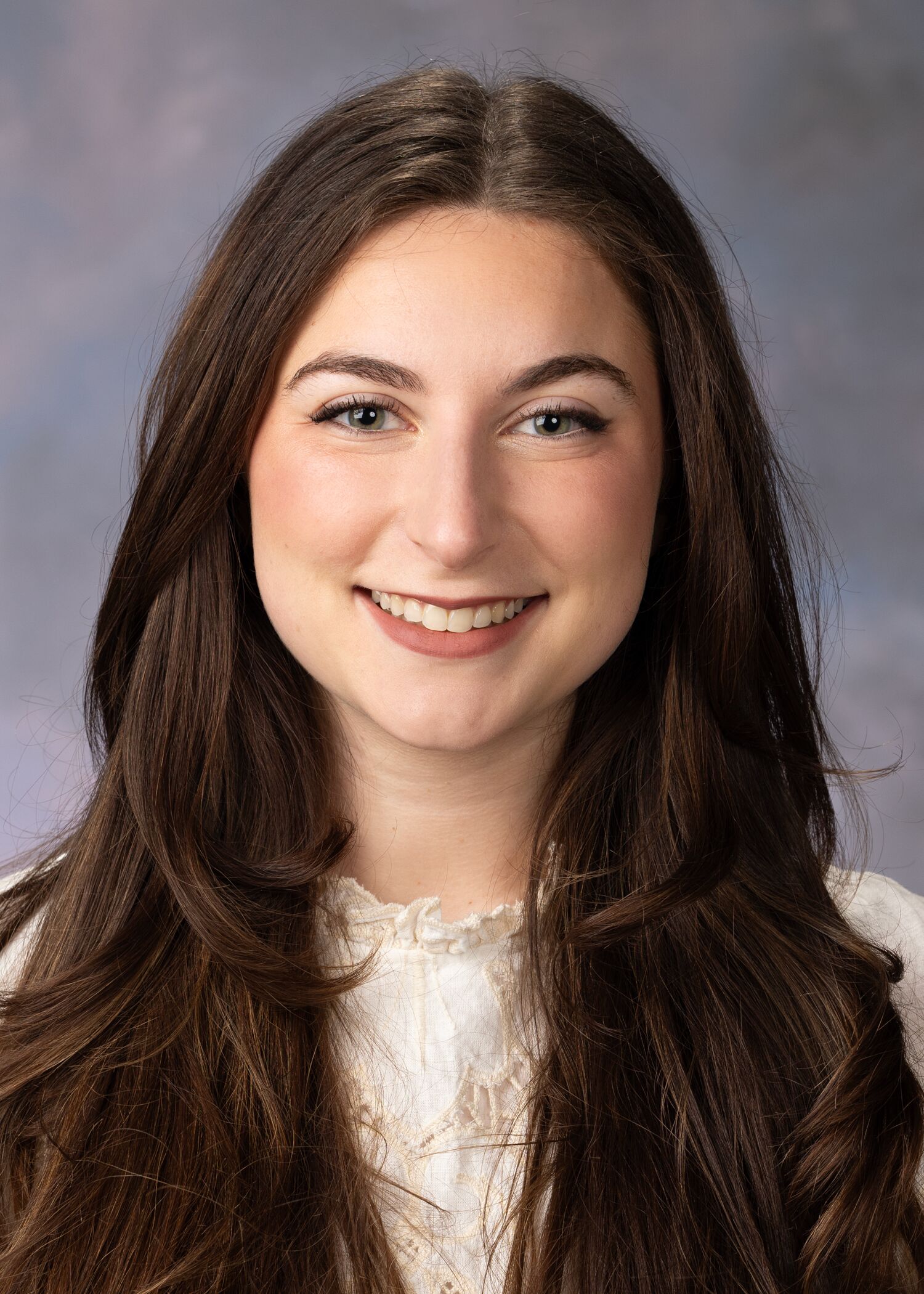 Portrait of Gabrielle Deutsch with long, dark brown hair wearing a white lace top, smiling at the camera. The background is softly blurred in shades of gray and purple.