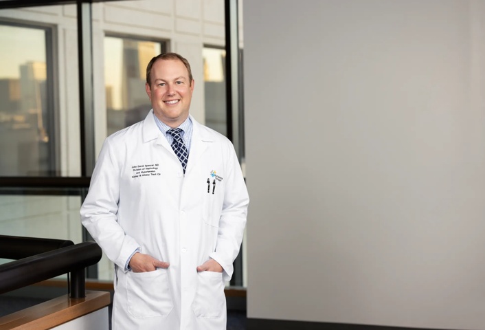 John Spencer is wearing a white coat with a shirt and tie underneath. He has his hands in his coat pockets and is smiling. Windows are shown in the hallway behind him.