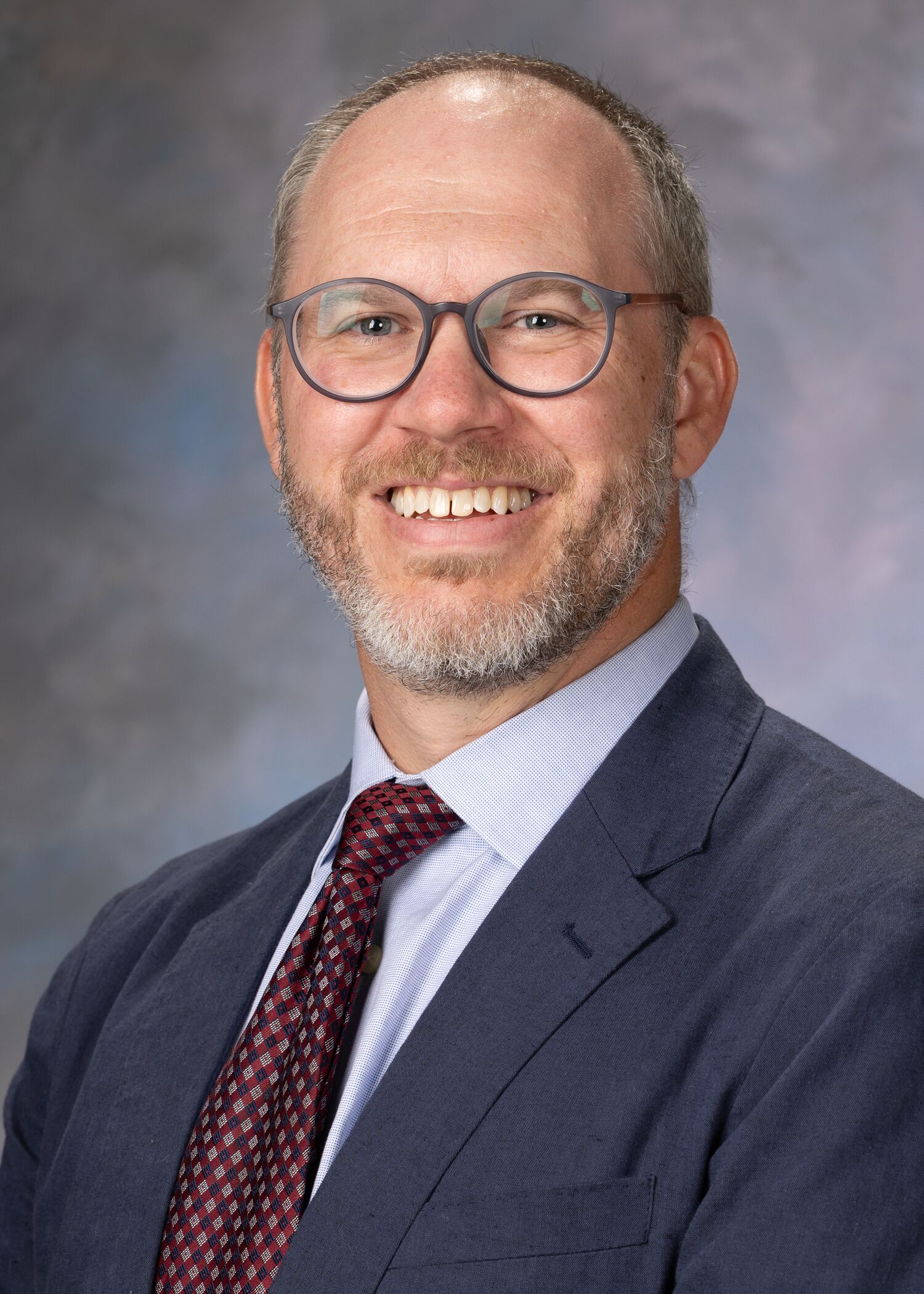 Portrait of Jon Campbell. Jon is wearing a navy blue suit jacket with a light blue button up and a maroon tie. He has glasses and a beard.