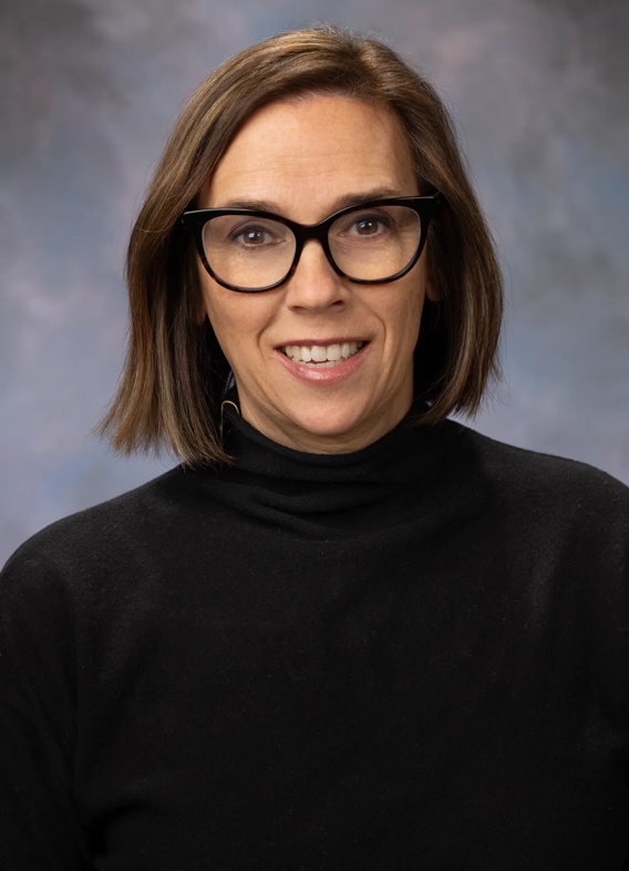 Portrait of Kendra Sauter, woman with short brown hair, glasses, and a black shirt.