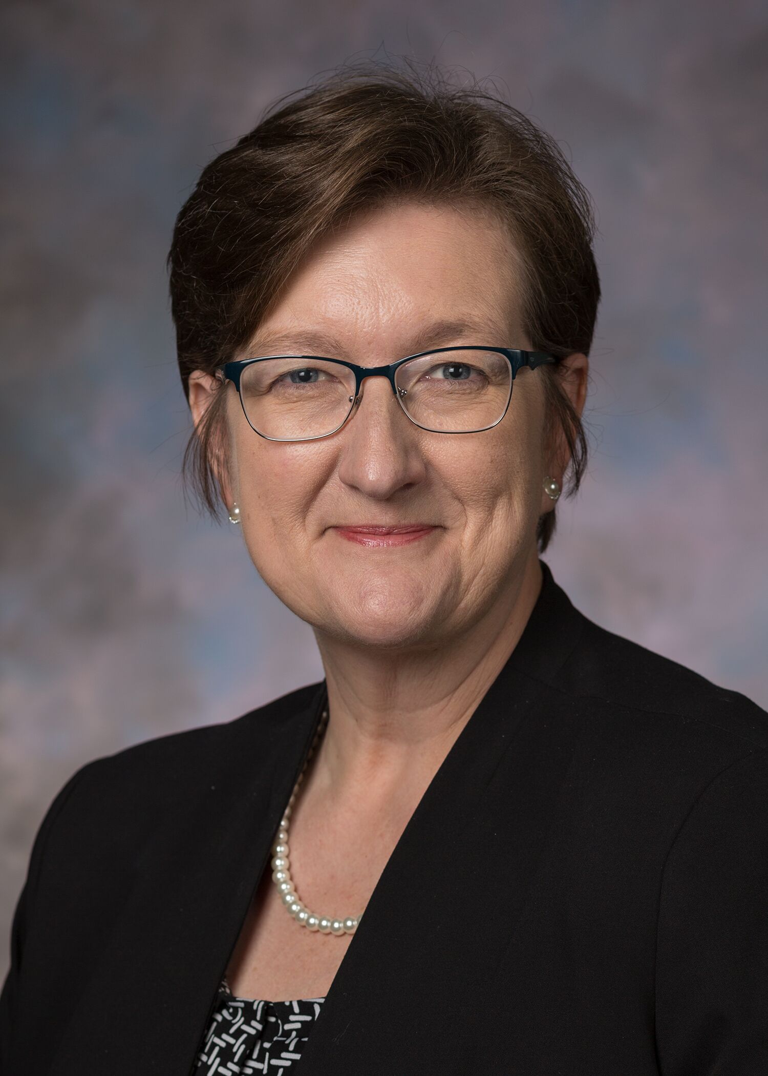 Portrait of Margie Brackeen. Margie has short brown hair, she's wearing glasses, a black jacket with a patterned top and a necklace. 